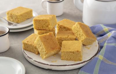 Foto da receita de Bolo de fubá sem glúten. Observa-se um prato de cerâmica com fatias quadradas do bolo e, ao lado esquerdo, uma xícara branca de café.