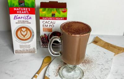 Foto da receita de chocolate quente cremoso servida em uma xícara alta e larga de vidro sobre uma tábua de mármore branca. Ao fundo há duas caixas de natures heart, uma de bebida vegetal barista e outra de cacau em pó