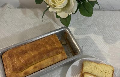 Fotografia vista de cima de um pão dentro de uma forma pequena retangular de alumínio ao lado de um prato pequeno raso com uma fatia do pão.