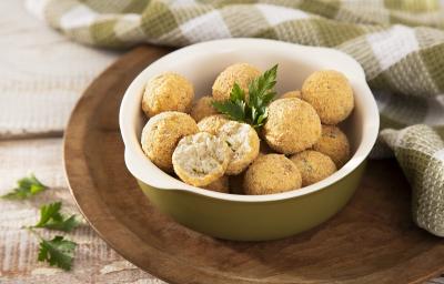 Foto em tons de bege da receita de bolinho proteico de frango servida em diversas porções dentro de uma panelinha de porcelana verde sobre uma tábua circular de madeira com um pano quadriculada verde musgo