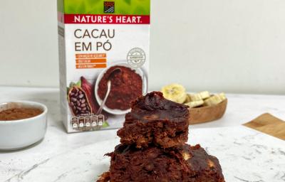 Foto em tons de marrom da receita de brownie de 3 ingredientes servida em diversas porções sobrepostas sobre uma de mármore branca. Ao fundo há um bowl com rodelas de banana e um com cacau em pó