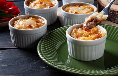 Fotografia em tons de marrom com vários ramekins ao centro. Dentro de cada recipiente existe uma receita de escondidinho de carne seca sem lactose. Um prato de mandiocas gratinadas com carne seca.