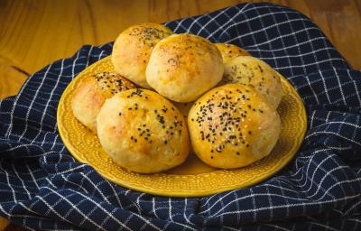 Foto aproximada de um prato amarelo com alguns pães de batata grande, decorados com gergelim. O prato está sobre um tecido azul quadriculado e numa bancada de madeira.