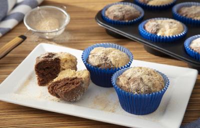 Foto da receita de muffin de cacau com farinha láctea. Observa-se dois muffins em forminhas azuis com farinha láctea polvilhada. Ao lado esquerdo, um muffin sem forminha cortado ao meio.