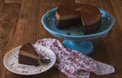 Foto da receita de bolo chocopudim da maria carpio, servido sobre um prato grande azul, com uma fatia cortada sobre outro prato branco, tudo em uma bancada de madeira