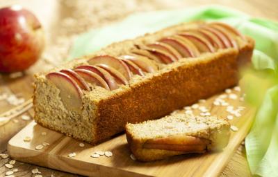 Fotografia em tons de marrom de uma bancada de madeira, sobre ela uma tábua de madeira com o bolo de maça. Ao fundo uma maçã e um paninho verde.