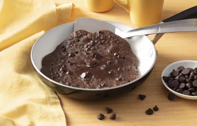 Fotografia em tons de amarelo em uma bancada amarela e um pano amarelo na lateral. Uma frigideira ao centro com o brownie de chocolate dentro dela. Ao lado, um potinho com gotas de chocolate.