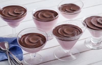 Fotografia em tons de chocolate e rosa, com seis taças contendo creme rosa e creme de chocolate, ao lado guardanapo azul e várias colheres, tudo sobre bancada em tom de rosa.