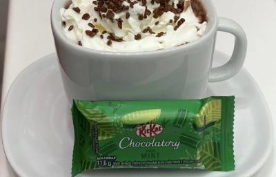 Fotografia de uma xícara branca com um hot chocolate com chantilly e granulado por cima. Na frente da xícara tem um kitkat de menta, e está sobre um prato pequeno de vidro raso branco.