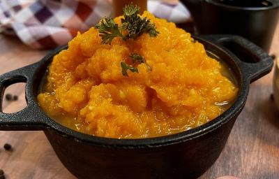 Foto em tons de laranja da receita de quibebe servida em uma panela de pedra preta com folhas salsinha para decorar. Ao fundo há um paninho quadriculado branco e vermelho além de uma xícara preta, tudo isso sobre uma mesa de madeira