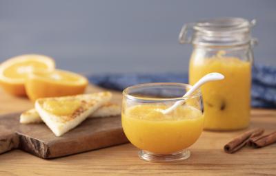 Fotografia em tons de laranja em uma bancada de madeira, uma tábua de madeira, um paninho azul, um pote hermético de vidro com geleia de laranja dentro. Ao lado, um potinho de vidro com mais geleia. Ao lado, fatias de pão e laranjas cortadas ao meio.