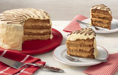 Fotografia em tons de vermelho em uma bancada cinza com paninhos vermelhos listrados e de bolinhas brancas. Ao centro, um prato branco com uma fatia do bolo de pão de mel com chocolate branco e à esquerda, um suporte grande vermelho com o bolo inteiro.