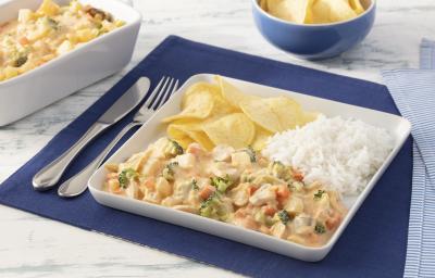 Fotografia em tons de azul em uma bancada de mármore branco com um jogo americano azul escuro, um prato quadrado branco grande com o strogonoff de legumes, arroz branco e batatas chips dentro dele. Ao fundo, um recipiente com mais strogonoff de legumes.