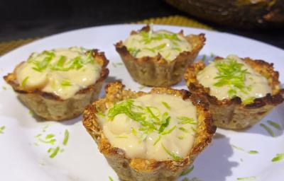 Foto aproximada de quatro mini tortinhas de limão, numa base em tom marrom claro e com um recheio cremoso, decoradas com raspas de limão em um prato branco