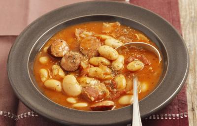 Fotografia de um prato fundo na cor preta com uma sopa de feijão branco e linguiça. Ao lado, três fatias de pão e do outro lado, um tempero em pó vermelho dentro de um pote pequeno branco de cerâmica, que está em cima de um pano em tons de vinho.