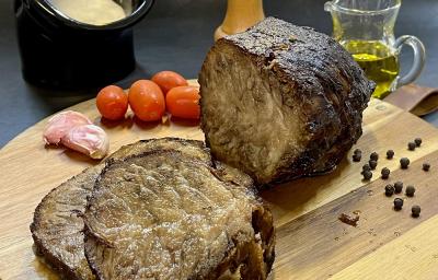 Foto em tons de marrom da receita de cupim na pressão servida em uma tábua de madeira com a peça de cupim cortada em algumas fatias e com tomates cerejas e pimentas. Ao fundo há uma jarra de vidro com azeite além de um recipiente preto com molho branco