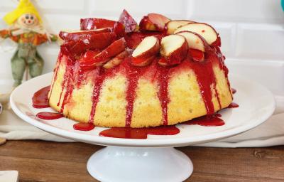 Foto da receita de bolo de maçã do amor, servido em um prato alto branco, com calda vermelha de maçã do amor escorrendo pelas laterais, com pedaços de maçã decorando o topo e itens de festa junina sobre a bancada ao lado do bolo