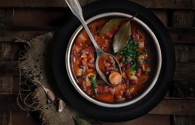 Fotografia vista de cima de uma sopa com linguiça, feijão, cenoura, costela e duas folhas de loura por cima. Apoiada na sopa, uma colher de sopa, que está dentro de um recipiente fundo de alumínio, sobre um prato preto, que está sobre uma mesa de madeira.