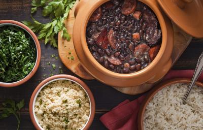 Fotografia em tons de preto e marrom de uma bancada de madeira escura, um pote com couve refogada, um pote com farofa, um pote com arroz branco e um pote com uma feijoada com feijão preto e várias carnes.