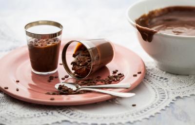 Foto em tons branco e rosa, ao centro prato rosa com dois copinhos contendo brigadeiro e duas colheres de sobremesa, ao lado pote com brigadeiro, sobre tolha branca em bancada de madeira.