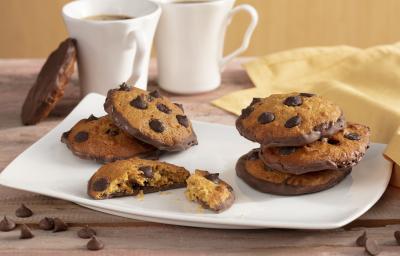 Fotografia em tons de amarelo em uma bancada de madeira com um pano amarelo, um recipiente quadrado branco com alguns cookies de cenoura recheado com chocolate. Ao fundo, duas xícaras brancas com café.