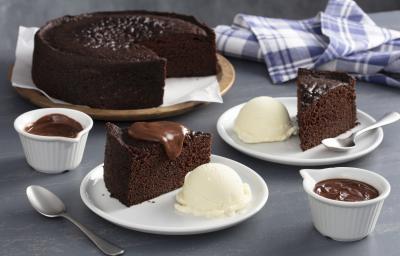 Fotografia em tons de marrom e azul de uma bancada cinza, ao centro dois pratos brancos com fatias de bolo de chocolate e ao lado uma bola de sorvete de creme. Ao fundo o bolo inteiro sem duas fatias.