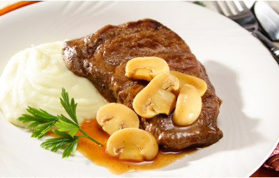 Fotografia em tons de branco e marrom, com bife com molho de champignons acompanhado de purê de batatas e ramo de salsa em prato branco, sobre sous plat marrom, ao lado guardanapo dobrado com garfo e faca por cima, tudo sobre toalha cinza.