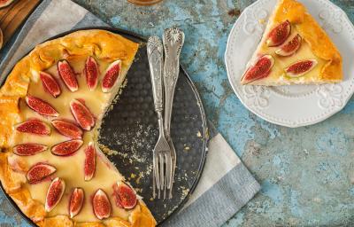 Fotografia vista de cima de uma torta de amêndoas com creme de leite e figos, coberta com figos fatiados, sobre o fundo falso da forma e uma fatia ao lado