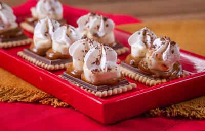 Fotografia em tons de vermelho com vários biscoitos Choco Biscuit ao centro. Por cima de cada biscoito existe uma camada de doce de leite, banana e, por cima, chantilly.