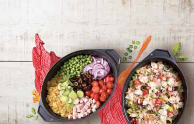 Fotografia em tons de cinza, vermelho, preto de uma bancada cinza vista de cima, contém um pano vermelho e por cima dois recipientes ovais um com macarrão, azeitona, peito de peru e tomates picados e no outro os mesmos ingredientes só que inteiros.