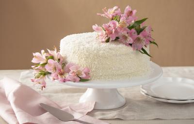 Foto da receita de Bolo de Gala. Observa-se um bolo grandioso com flores rosas sobre uma boleira de cerâmica branca. Atrás, um fundo nude e pratinhos de sobremesa decoram a foto, assim como um guardanapo rosa.