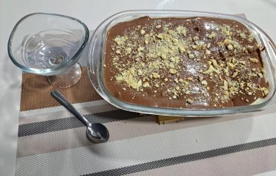 Foto da receita de Pavê de Cupuaçu com Ganache de Chocolate. Observa-se uma travessa retangular com o doce decorado de biscoito picado por cima e ao lado esquerdo tem uma taça individual.