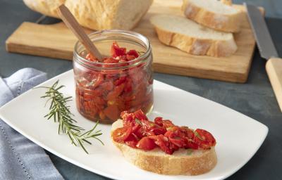 Foto da receita de Tomate confit. Observa-se uma torrada com os tomates assados em cima e um recipiente transparente de vidro com os tomates armazenados. Alecrim e pedaços de pão fatiados decoram a foto.