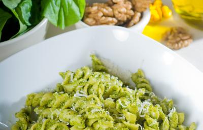 Fotografia em tom claro e branco, com um prato quadrado fundo branco no centro da foto, dentro tem macarrão no estilo parafuso com pesto em tom verde claro e queijo ralado por cima. No fundo da foto tem manjericão e pinolis, tudo sobre uma mesa branca..