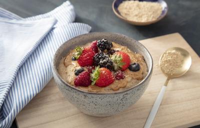 Foto em tons de bege da receita de mingau nutritivo servida em um bowl de cerâmica sobre uma tábua de madeira clara com uma colher prateada ao lado. Ao lado ainda há um paninho azul listrado e um recipiente com aveia em flocos