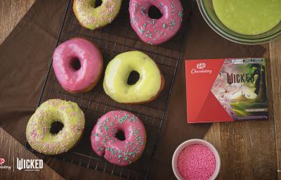 Foto da receita de Donuts Wicked, vistos de cima, servidos em uma grade de ferro preta. São seis no total, decorados com cobertura rosa e verde e alguns com granulados coloridos. Ao lado direito há uma caixa de KitKat Wicked.