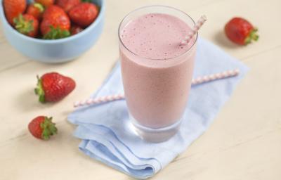 Fotografia em tons de rosa e azul de uma bancada rosa com um paninho azul, sobre ele um copo de vitamina com um canudo. Ao lado morangos. Ao fundo um recipiente redondo azul com morangos.