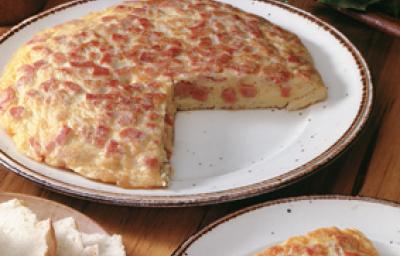 Fotografia em tons de vermelho em uma mesa de madeira com um prato branco raso grande com a omelete cortada ao meio. Ao lado, um prato com a fatia da omelete e outro prato com fatias de pão.