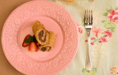 Fotografia em tons de rosa em uma mesa de madeira, uma toalha branca com flores rosas, um prato redondo raso rosa com os rolinhos recheados de chocolate e morango. Ao lado, um garfo.