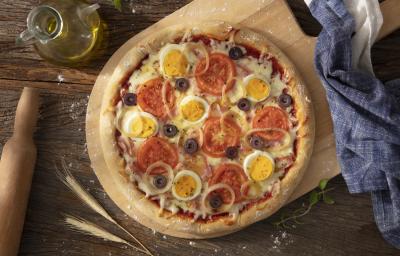 Fotografia em tons de azul em uma bancada de madeira escura, um paninho azul, uma tábua de madeira com a pizza portuguesa em cima dela. Ao lado, um potinho de azeite e um rolo de massa.