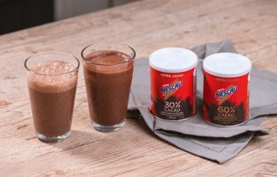 Foto da receita de smoothie pós treino de nescau extra cacau servida em dois copos de vidro altos sobre uma mesa de madeira com duas embalagens de nescau extra cacau 30% e 60% sobre um paninho cinza