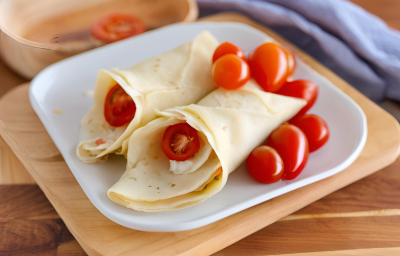 Fotografia em tons de bege e azul com um prato branco ao centro. Dentro do prato existe dois enrolados de crepe recheados com creme de palmito com NINHO Primeira Infância e tomates cereja.