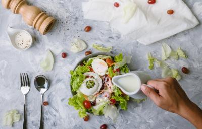 Fotografia vista de cima de uma mão segurando uma jarra pequena na cor branca, que está caindo um molho branco em uma salada de folhas, cebola, ovo cozido, maçã, tomates cereja, cenoura e azeitona preta. O refratário está sobre uma mesa de mármore branca.