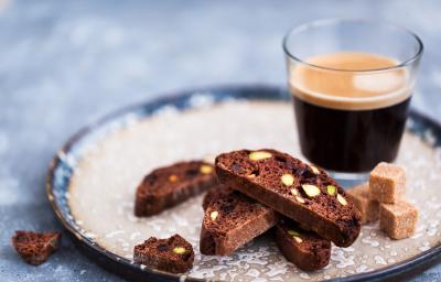 Fotografia quatro fatias de um torrão de chocolate ao lado de cubinhos de açúcar, para acompanhar um Nescafé Espresso Intenso que está em uma xícara de vidro, sobre um prato de vidro desenhado, de cora branca e azul.