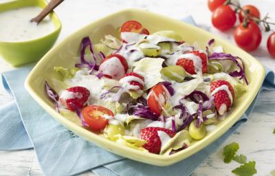 Fotografia em tons de branco, azul e verde de uma bancada branca e um paninho azul, sobre ele um recipiente quadrado verde com a salada e o molho. Ao fundo um recipiente quadrado verde com o molho, uma colher e tomatinhos cereja.