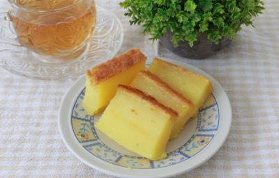 Foto da receita do Bolo de Macaxeira Cremoso. Observa-se um prato de sobremesa com quatro fatias finas do bolo cremoso. Ao lado esquerdo, uma xícara transparente de chá sobre uma toalha quadriculada off white