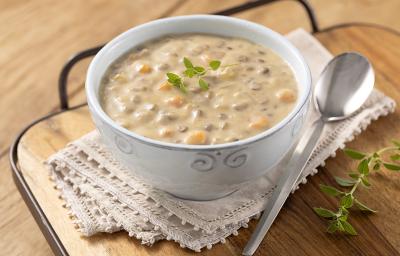 Foto em tons de amarelo com a receita de creme de lentilha, requeijão NESTLÉ e peito de peru servida em um bowl branco com adornos sobre um pano de renda bege em cima de uma tábua de madeira e uma colher prateada à frente. Tudo em cima da mesa de madeira.