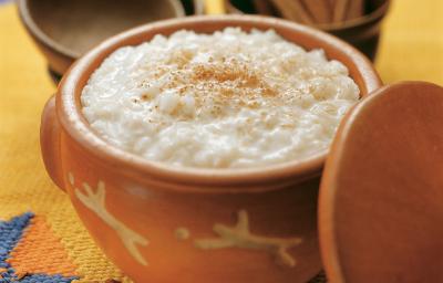 Fotografia em tons de amarelo em uma mesa de madeira com uma toalha amarela desenhada, um recipiente de barro com o arroz doce dentro dele. Ao fundo, um potinho com paus de canela.