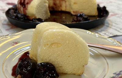 Foto da Receita de Pudim de Claras. Observa-se um prato de sobremesa branco de porcelana com uma fatia do pudim e calda de ameixas. Atrás, o pudim inteiro com a calda.