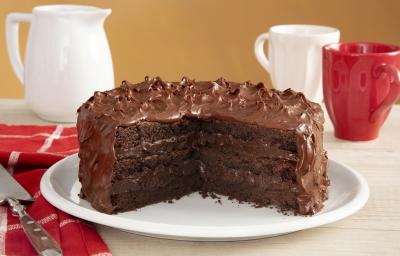 Fotografia em tons de vermelho em uma bancada de madeira com um pano vermelho listrado em branco, com um prato branco no centro com o bolo brownie com brigadeiro de cacau de cobertura. Ao fundo, uma xícara branca, uma xícara vermelha e um bule branco.